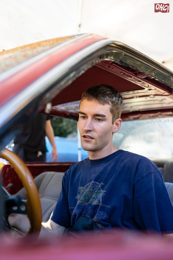 Young man in a blue shirt, sitting inside a red car under repair, with OHG! logo in the top right corner.