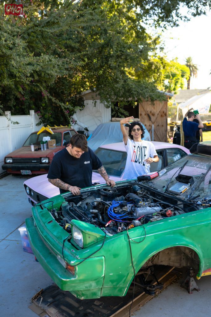Two men working on a green car's engine with a pink car in the background in an outdoor setting, one waving.