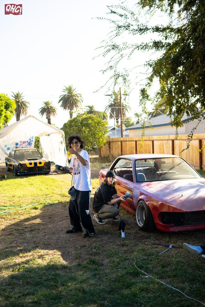 Two people working on a red car outdoors, one using a tool, and another pointing, with a yellow car in the background.
