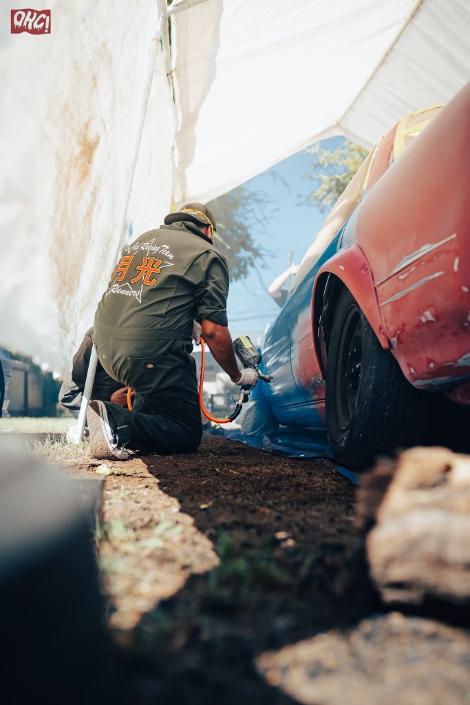 Mechanic painting a car under a tent, wearing a jacket with 月光 Runners (Moonlight Runners) written on the back.