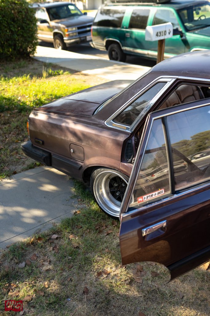 A vintage car with an open door, displaying a sticker that reads トヨタパート 8633 (Toyota Part 8633), parked on a sunny street.