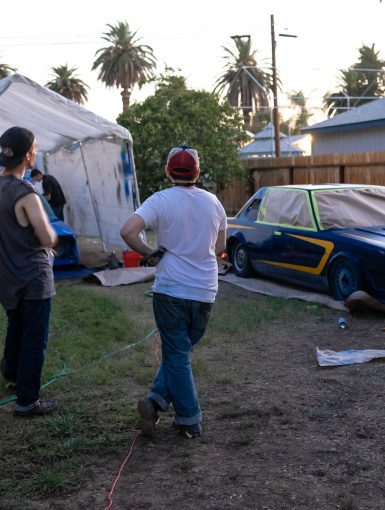 People painting custom race car outdoors in a backyard garage setup with tarp shelter and painting equipment.