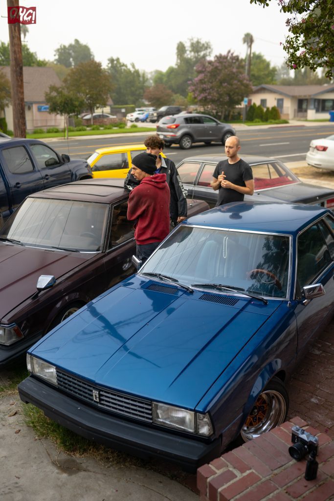 Group of people standing beside classic parked cars on a suburban street; vintage camera visible on brick ledge.