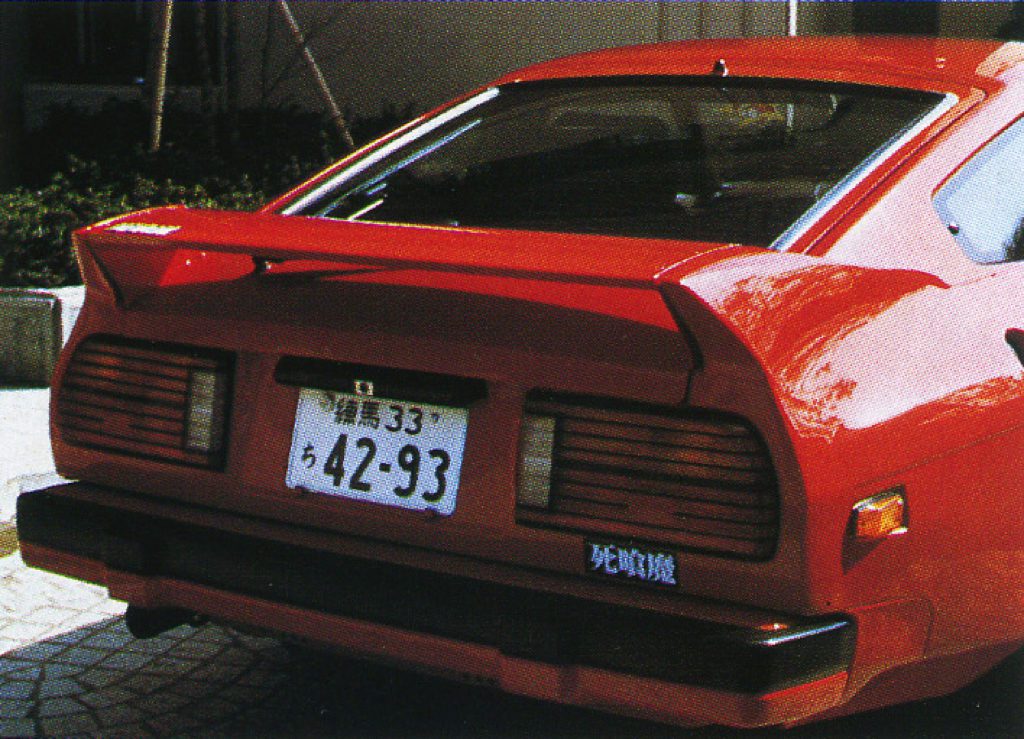 Rear view of a red sports car with a Japanese license plate and a 'Battle Machine' decal parked on a sunny street.
