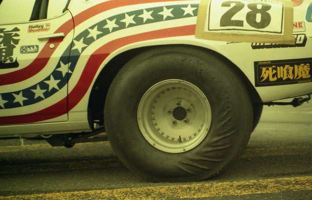 Close-up of vintage race car with number 28, American flag design, and Japanese Death Machine decal, parked on race track.