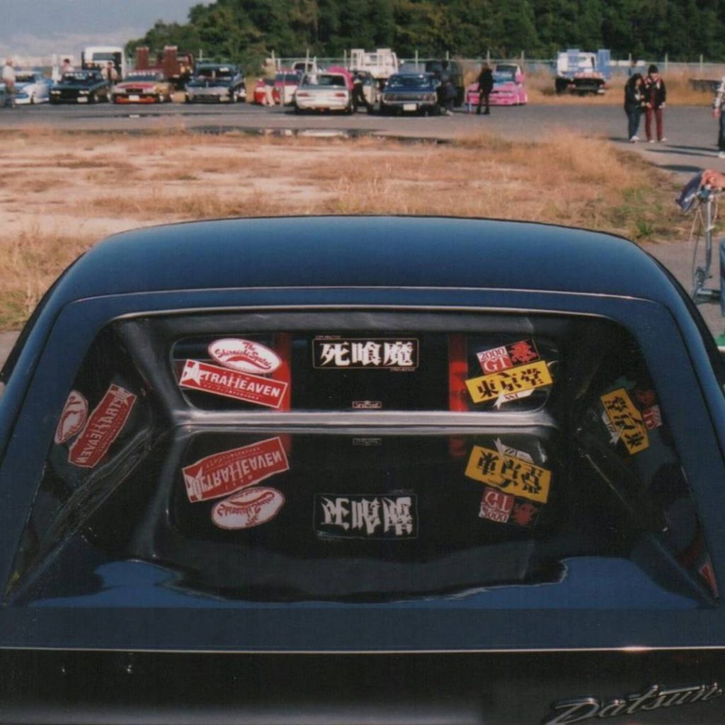 Rear view of a car with various Japanese car stickers on the back window, showcasing custom decals at an outdoor car meet.