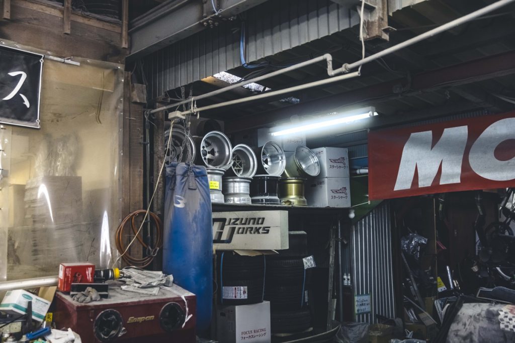 A cluttered garage with car wheels, tires, boxes, tools, and a hanging punching bag, under dim fluorescent lighting.