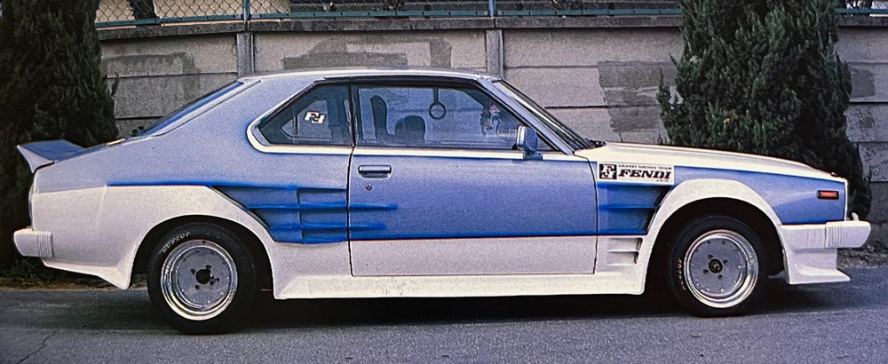 Custom blue and white retro car with Fendi logo parked on street next to concrete wall and trees.