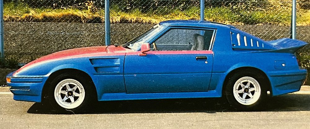A modified blue and red Mazda RX-7 sports car is parked in front of a fence on a sunny day.