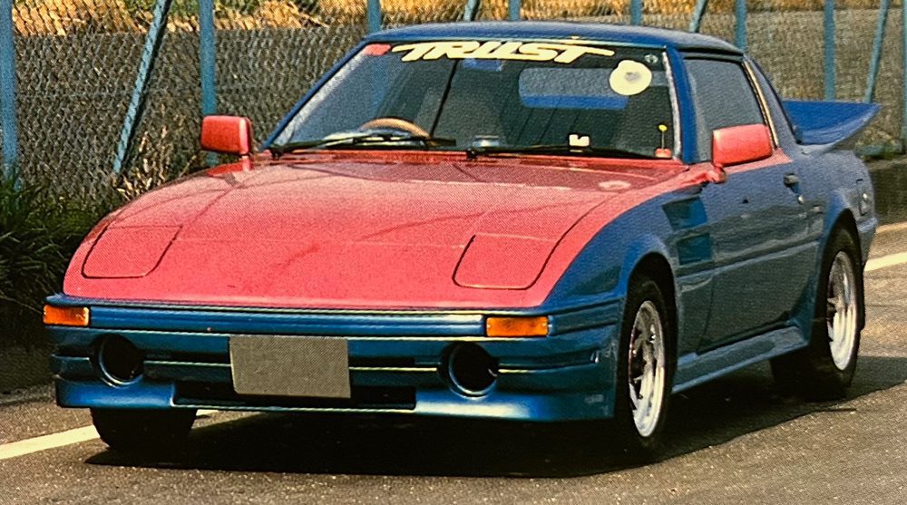 Modified vintage Japanese sports car with TRUST windshield decal, red and blue paint, parked on a sunny road.