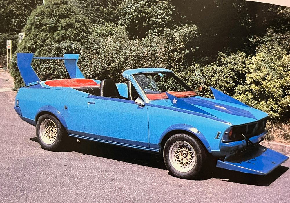 Vibrant blue customized Japanese car with large wing spoilers, parked on a street beside lush green trees.
