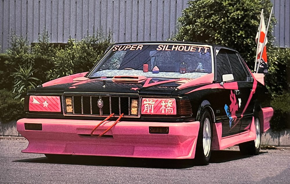 Custom black and pink car with SUPER SILHOUETT text, Japanese decals, and unique body kit, parked by greenery.