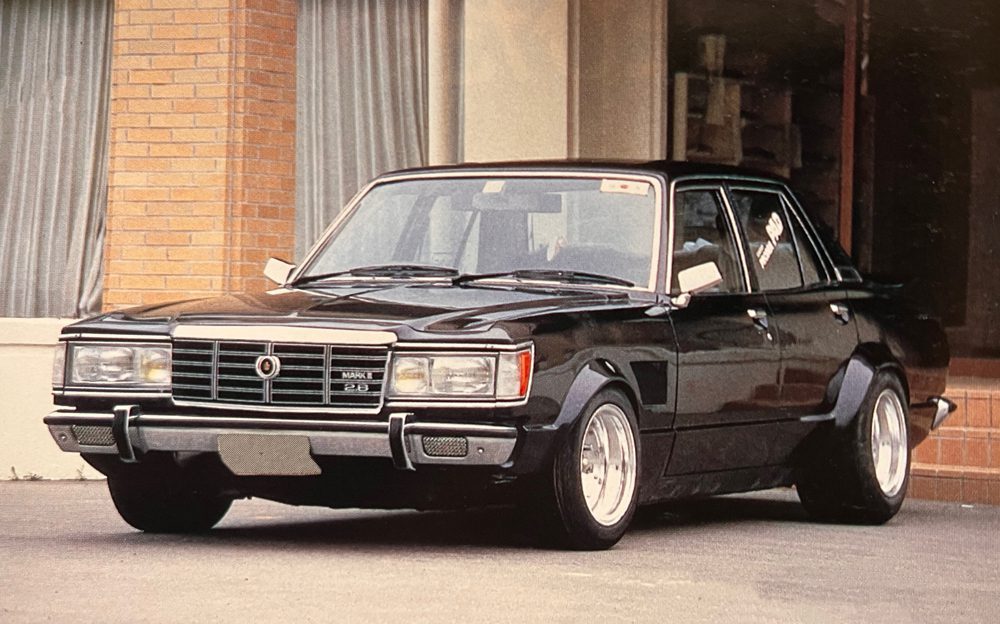 Classic black Toyota Mark II hardtop parked in front of a building. Model number 26 visible on the grille.
