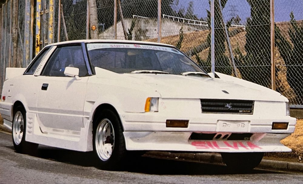 White modified Nissan Silvia parked on the street, showcasing front-lip spoiler and custom decals in Japanese text.