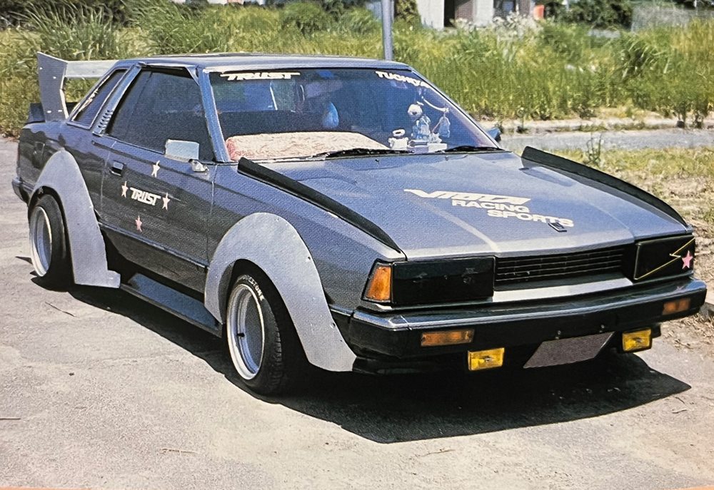 Modified vintage car with custom body kit and TRUST decals, parked outdoors on a sunny day.