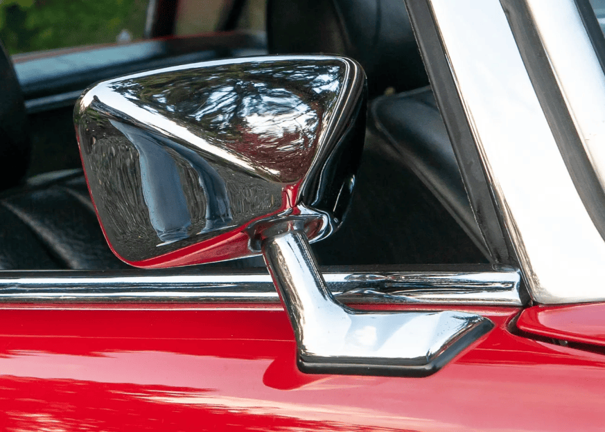 Close-up of a vintage car's chrome side mirror on a red door, reflecting a scenic outdoor view.