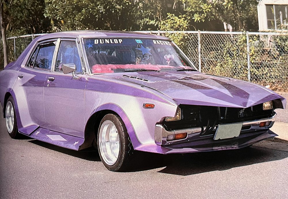 Modified purple vintage car with aerodynamic body kit and DUNLOP and RACING decals on windshield, parked by a fence.