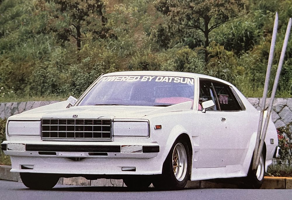White retro car with Powered by Datsun windshield decal, parked outdoors in front of green trees and stone wall.