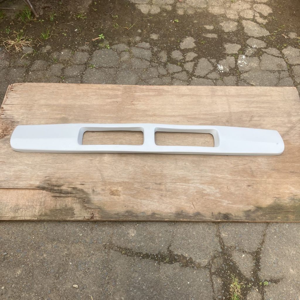 White painted car bumper with dual openings placed on wooden surface. Background shows a stone pavement.