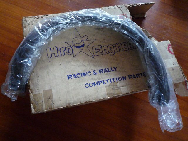 Box labeled Hiro Engineer Racing & Rally Competition Parts with a curved part wrapped in plastic on a wooden floor.