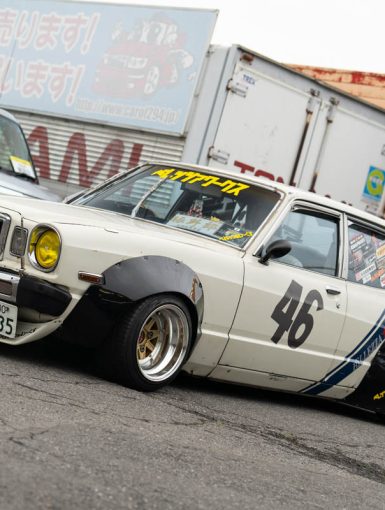 Classic Japanese car with Yokohama Advan livery, parked by a garage; racing number 46; yellow headlights; retro-mod style.