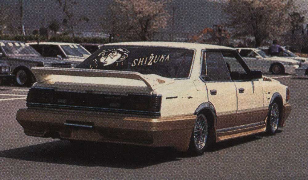 Retro modified car with large rear spoiler in a Japanese parking lot, featuring Shizuka artwork on the back window.