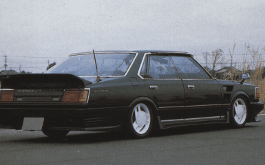 A black vintage Nissan Gloria sedan with a spoiler, low stance, and white wheels on a deserted road.