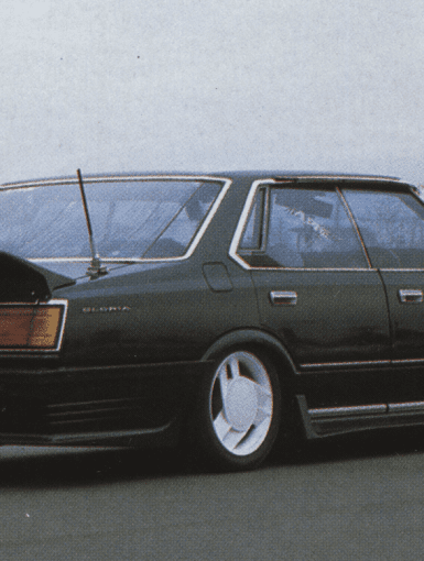 A black vintage Nissan Gloria sedan with a spoiler, low stance, and white wheels on a deserted road.