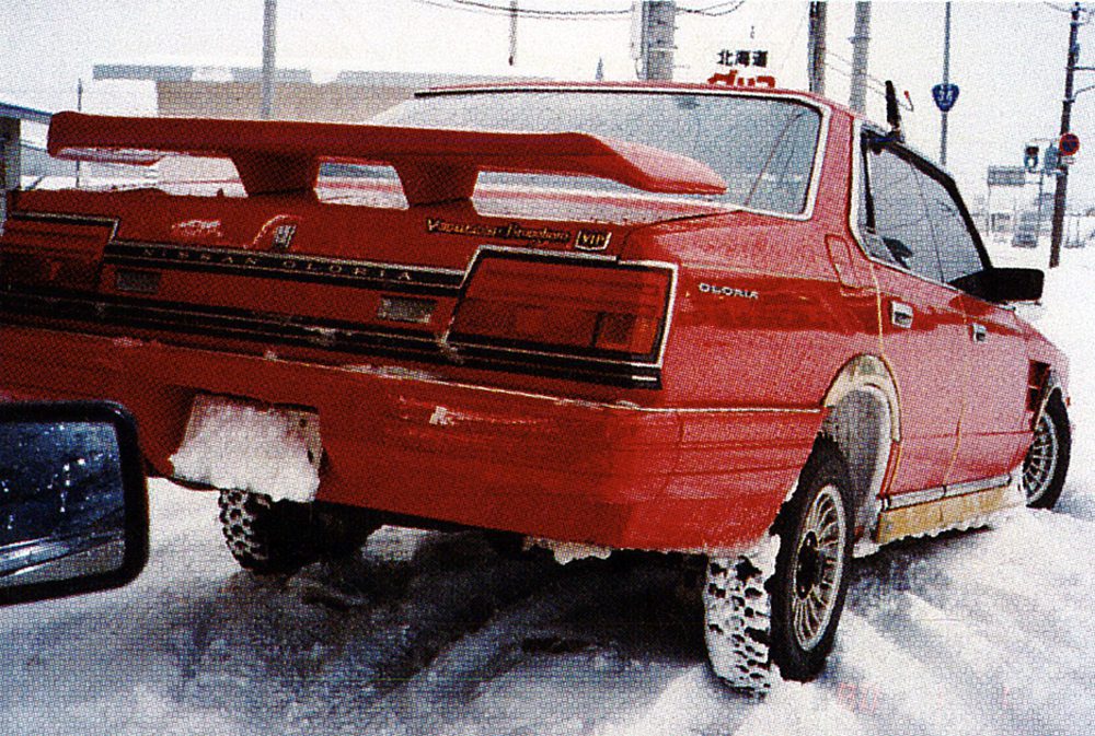 Red vintage car with snow tires driving through snowy streets.