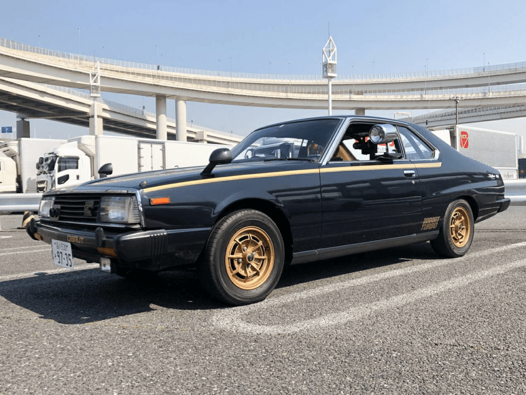 Vintage black car parked under a highway bridge, showcasing classic design and golden wheels.