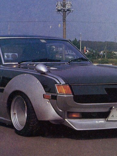 Vintage modified car with wide fenders and sleek design parked on a road with landscape in background.