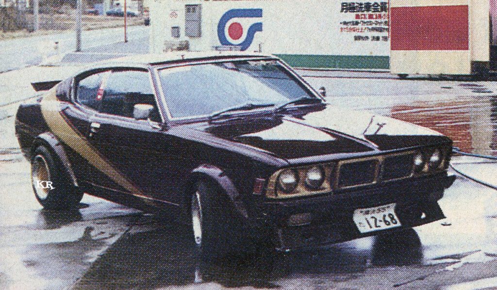 Vintage sports car parked at a gas station, showcasing a retro design with a dark exterior and stylized fenders.