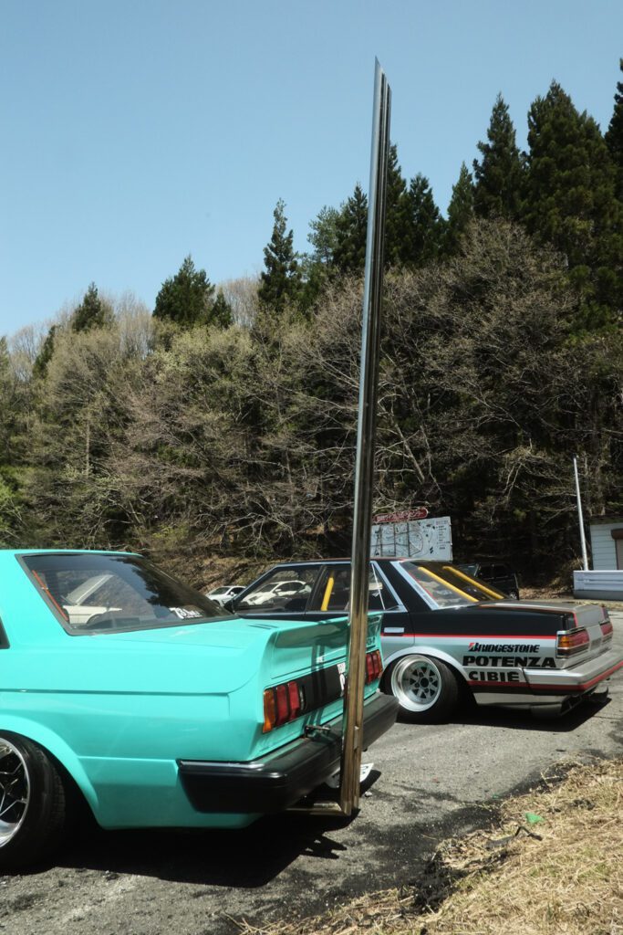 Two modified vintage cars parked outdoors with a tall exhaust pipe, surrounded by trees under a clear sky.