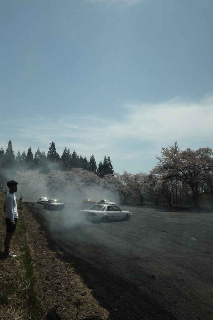 Cars drifting on asphalt amid cherry blossoms, emitting smoke under clear blue sky.