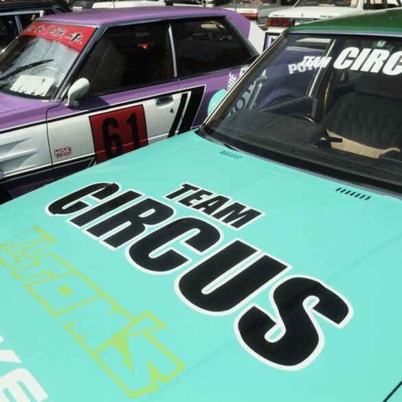 Vintage cars with Team Circus logos, featuring teal and purple designs, parked side by side.
