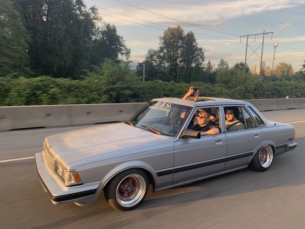 Vintage car cruising on a highway with passengers capturing the moment. Trees and a clear blue sky in the background.