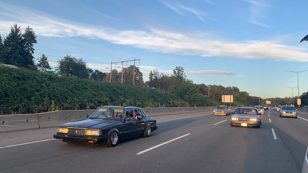 Cars driving on a sunny highway with green trees and a blue sky in the background.