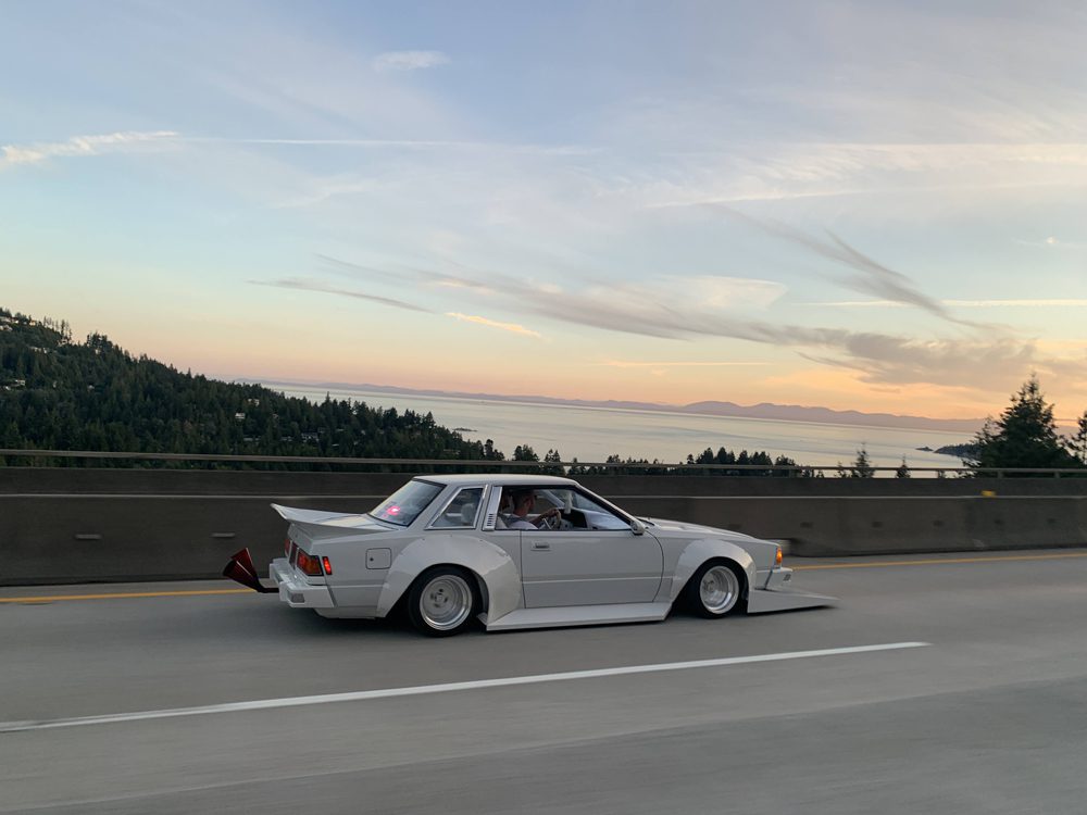 White modified car cruising on a scenic highway at sunset, with ocean and forested hills in the background.