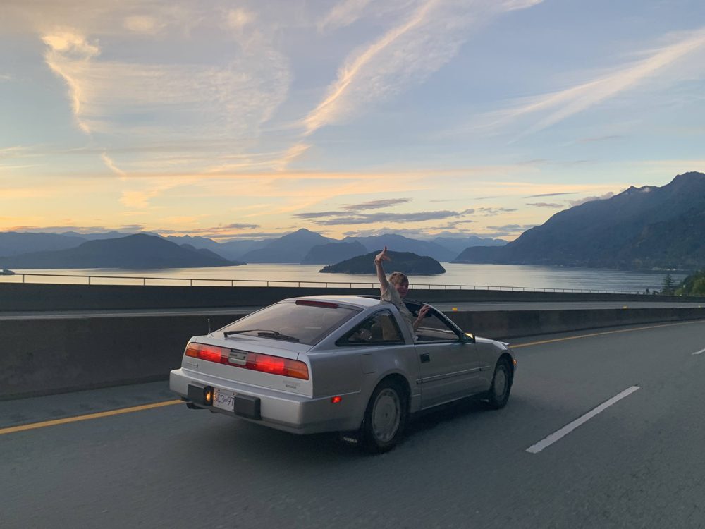 Classic silver sports car on scenic lakeside highway at sunset with mountains in background.
