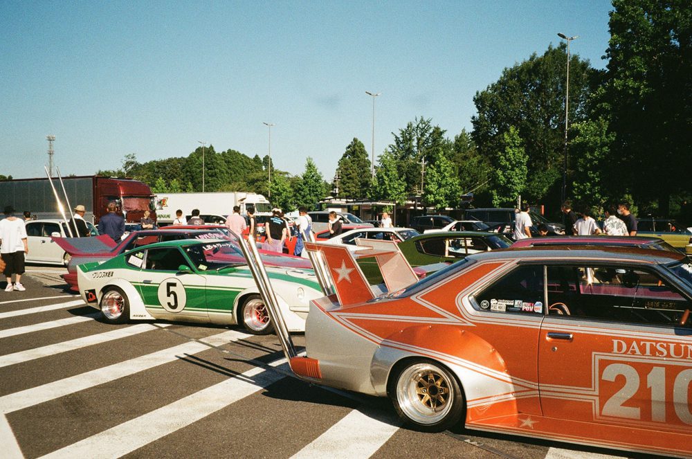 Vintage cars at a car show, featuring customized paint and spoilers, with a crowd and trees in the background.