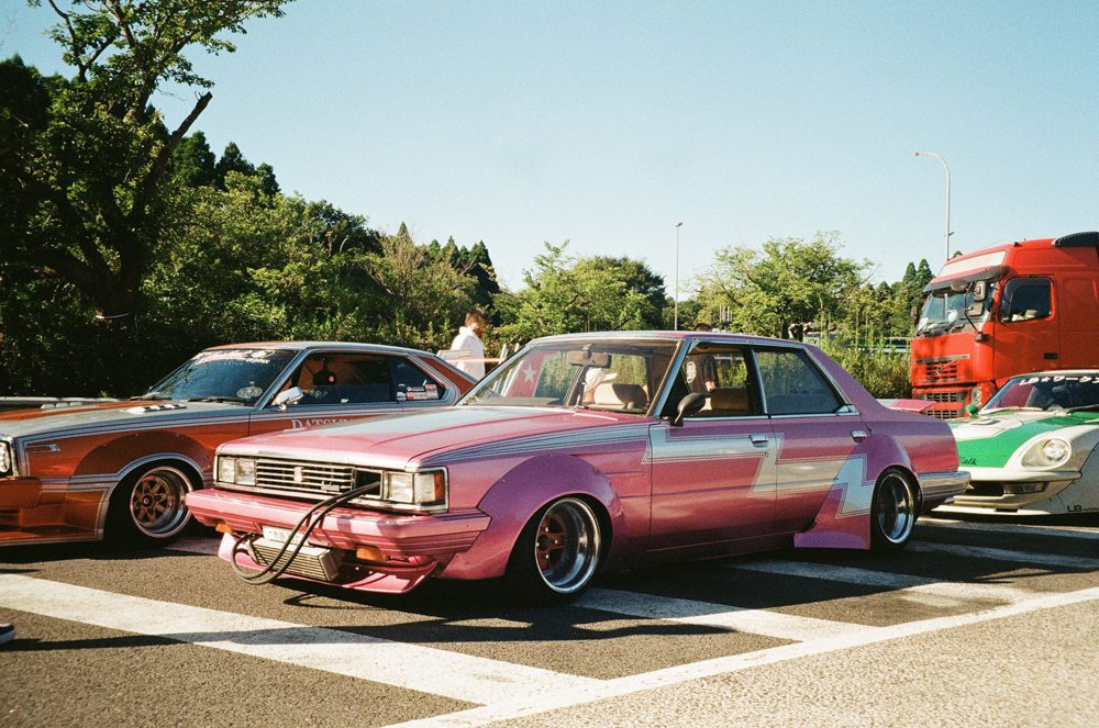 Custom pink vintage car with unique mods at a car meet, parked between other modified vehicles on a sunny day.