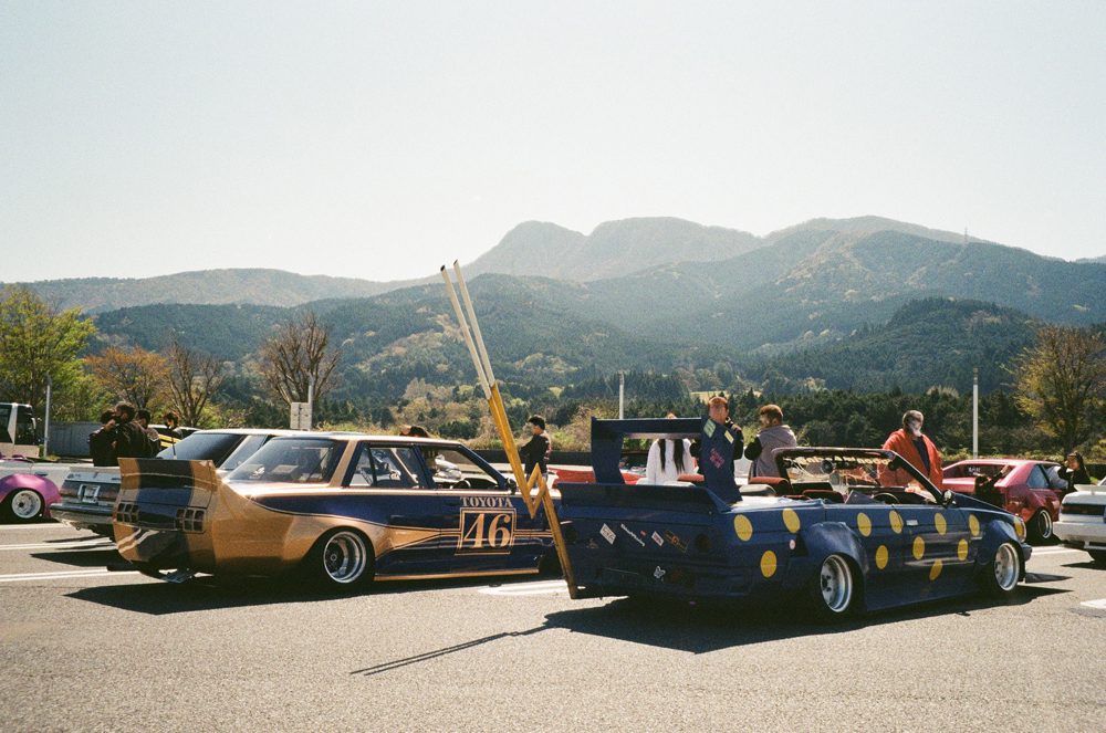 Vintage Japanese cars at a car meet with mountain backdrop and clear skies.