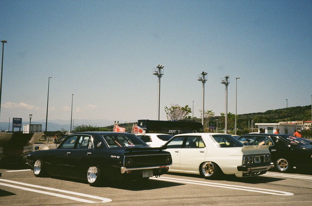 Classic cars parked outdoors at a car meet under a clear sky, mountains visible in the background.