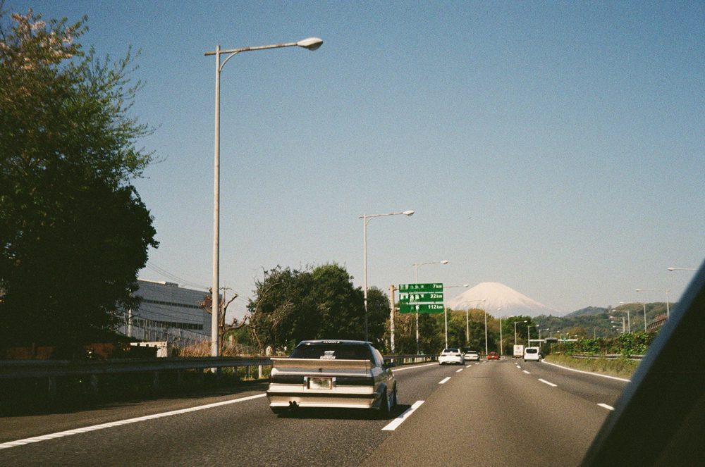 Cars driving on a highway towards Mount Fuji, with clear blue skies and road signs visible.