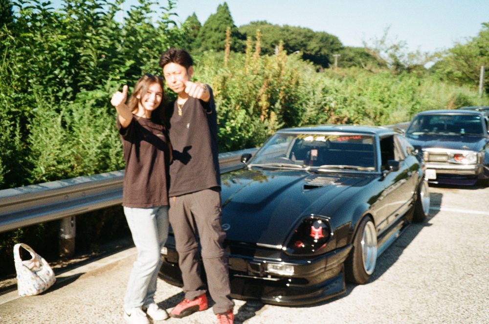 Two people posing happily with a sleek black car on a sunny day, surrounded by lush greenery.