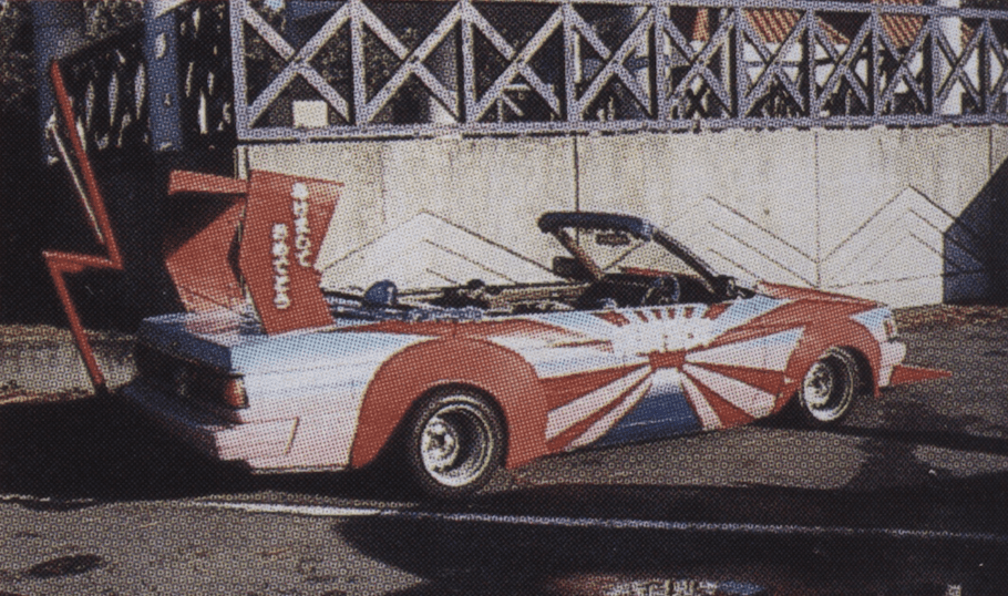 Custom car with red, white, and blue graphics, large rear spoiler, and open top in a sunny outdoor setting.