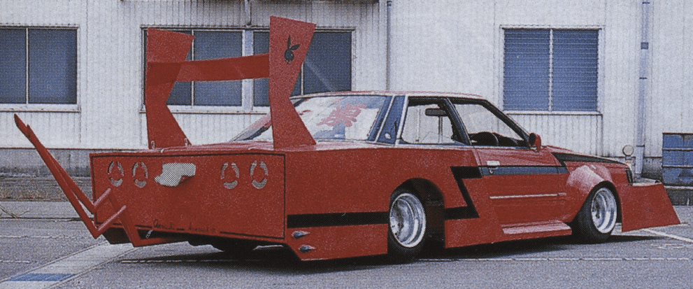 Red custom car with large rear spoiler and unique body kit, parked in front of a building.