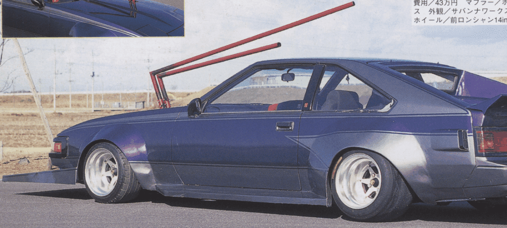 Modified vintage car with large rear wing and unique exhaust pipes in a scenic outdoor setting.