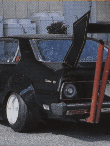 Modified classic black car with large rear wing and dual exhaust pipes for a unique street racing style.