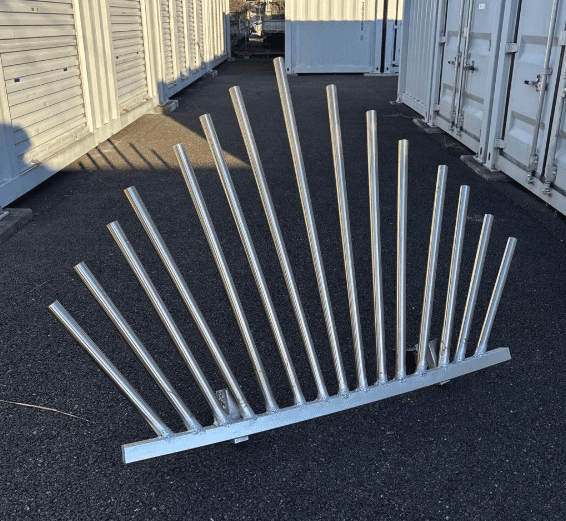 Metallic sculpture with vertical rods on asphalt, flanked by shipping containers in industrial setting.